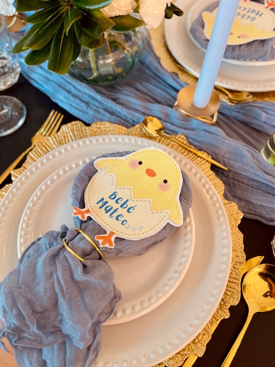 Mesa de baby shower con base dorada, servilleta gris y tarjeta de pollito
