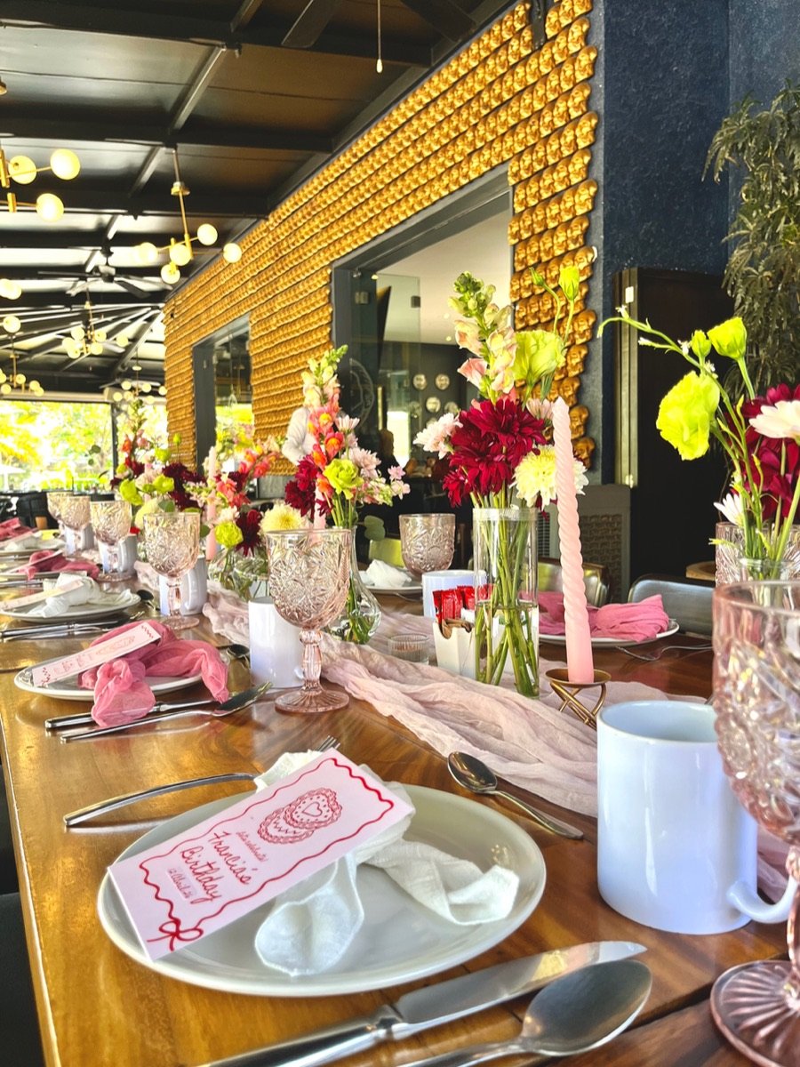 Mesa larga montada en restaurante, con copas talladas, velas rosas y centros de flores